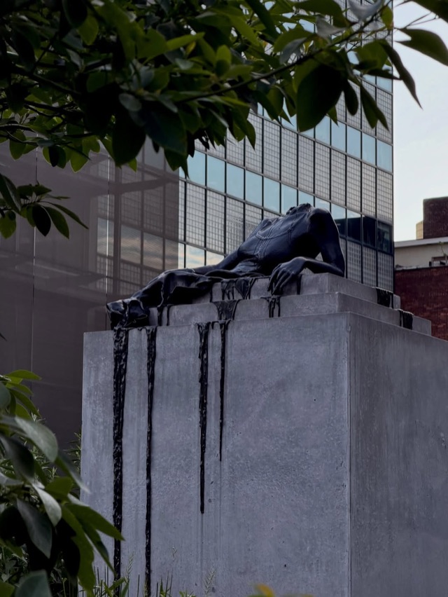 Dark bronze figure draped over a concrete plinth, framed by leaves
