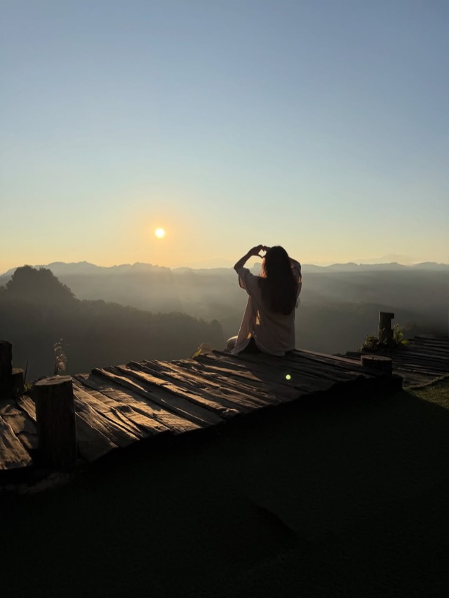 A figure making a heart with her hands toward a hazy mountain sunrise